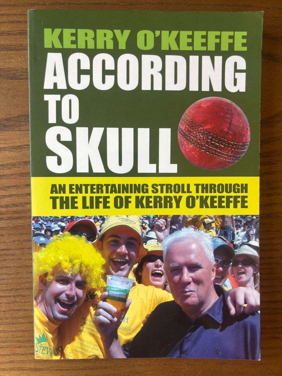 The book cover of 'According to Skull' by Kerry O'Keeffe, featuring a dark green top half with bold white and lime green text, a red cricket ball graphic, and a yellow band with black text above a photo of cheering fans in yellow clothing.