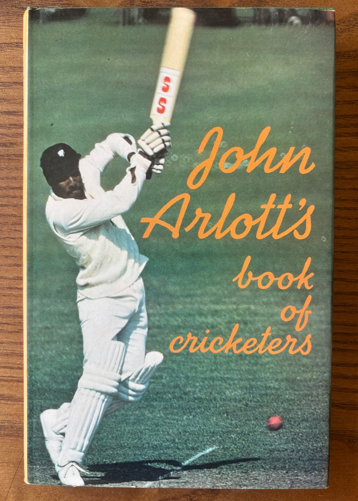 The front cover of the book 'John Arlott's book of cricketers' resting on a wooden surface. The cover features a vintage photograph of a cricket player in white uniform mid-swing, with the title written in orange script.