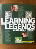 The front cover of the book 'Learning Australian Cricket' by John Buchanan, featuring a dark green background with large white title text and multiple inset photos of cricket players, photographed on a wooden surface.