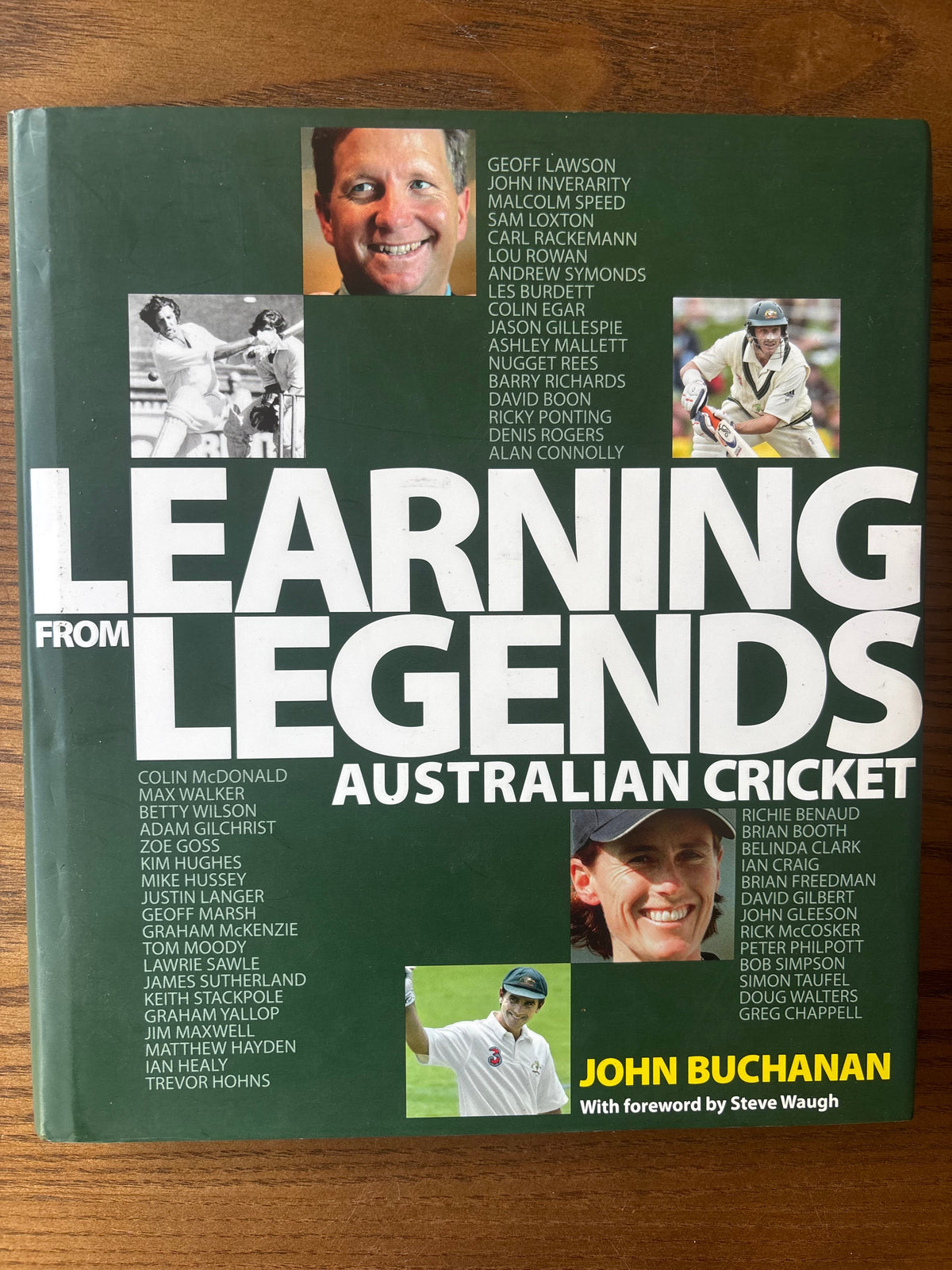 The front cover of the book 'Learning Australian Cricket' by John Buchanan, featuring a dark green background with large white title text and multiple inset photos of cricket players, photographed on a wooden surface.