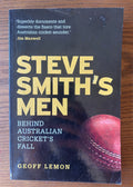 The front cover of the book 'Steve Smith's Men' by Geoff Lemon, featuring a black background with gold and white text and a red cricket ball graphic, photographed on a wooden surface.
