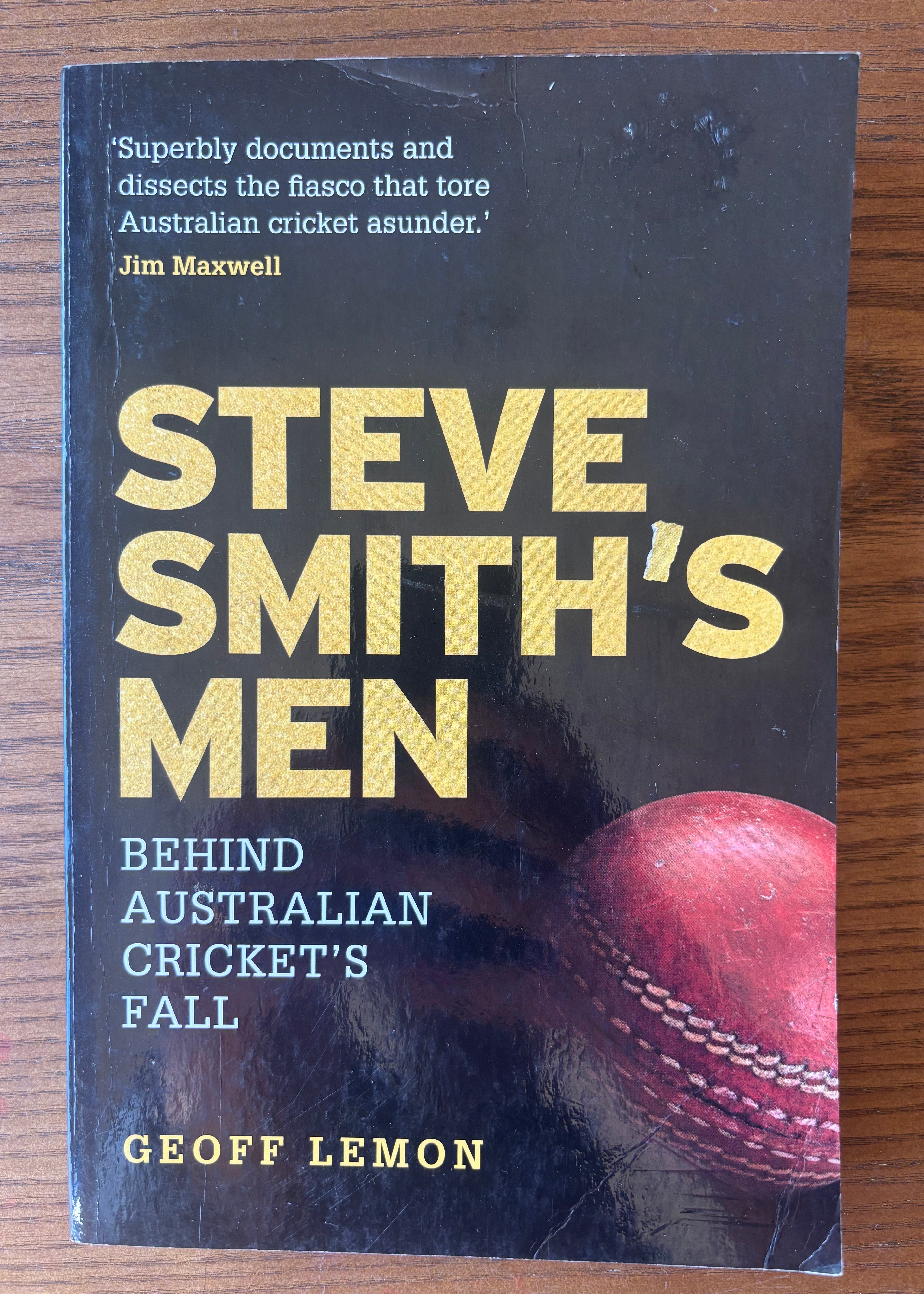 The front cover of the book 'Steve Smith's Men' by Geoff Lemon, featuring a black background with gold and white text and a red cricket ball graphic, photographed on a wooden surface.